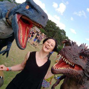 A photo of Anna Smith Spark - a white woman with short black hair, wearing a black v-neck dress. There are dinosaur statues on either side of her.