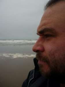 Side on close up image of a man's face as he looks away from the camera. He has dark hair and beard, and is on a beach.