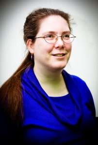 Image of a young woman looking off to the side of the camera. She is white, has long brown hair held back in a ponytail, and is wearing glasses and a blue cowl neck top