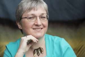 A photo of Juliet E McKenna - a white woman with short, greying hair, wearing a turquoise top and a bold necklace with three large metal feathers hanging from it. 