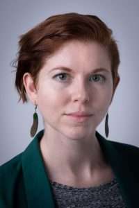 Photo of L.R. Lam, a young-looking white person with femme presentation. Lam has short red hair, a blue-grey top and green jacket. They are wearing feather earrings