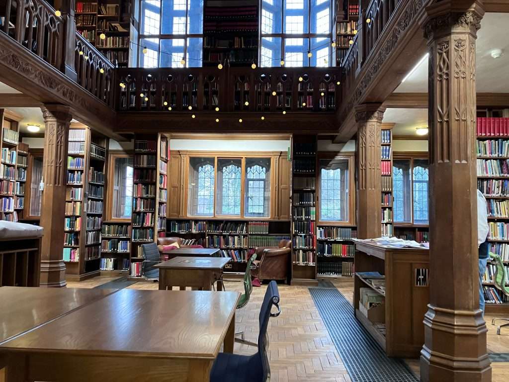 An interior shot of the library. Desks line the middle of the room while the walls are covered in bookcases. Wooden pillars hold up the first-floor balcony. Fairy lights are draped from the balcony
