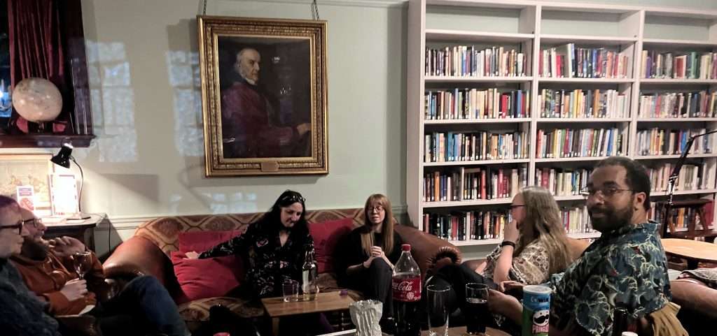A group of people sitting on sofas and armchairs beside a large bookcase. They are talking and laughing.