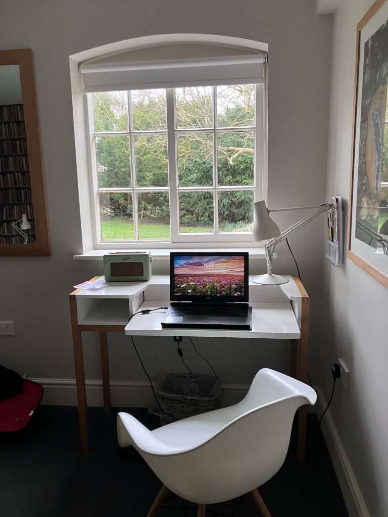 A white desk situated in front of a window looking out on a garden. There are trees and bushes visible through the window. A laptop, radio and lamp all sit on the desk.