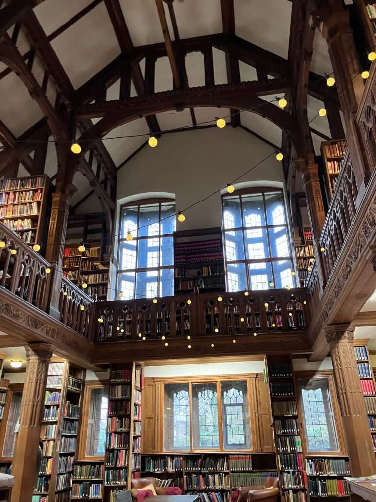 Interior shot of the library, looking up towards the high ceiling lined with wooden beans. Large bow windows dominate one wall while rows of fully-laden bookcases can be seen to either side.