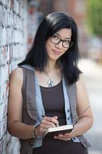 A dark-haired woman in glasses leans against a wall and looks towards the camera, smiling