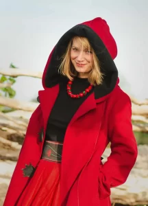 Gabriela Houston - A fair haired woman in a long red coat and hood smiles at the camera.