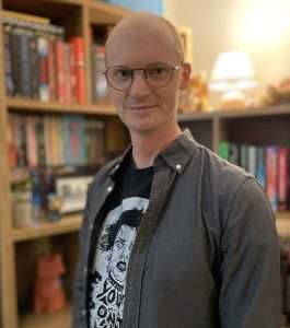 A fair-haired man in glasses looks towards the camera. There are bookcases behind him
