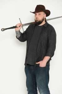 Lee C Conley - A man wearing a stetson  is holding a sword over his shoulder and looking towards the camera in a side-on pose