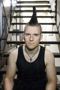 A man with a mohawk, wearing a dark vest top, sitting on stairs and looking at the camera