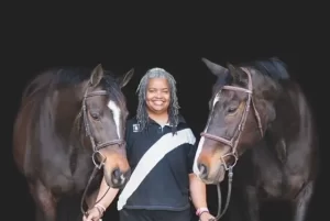 A woman in dark clothing stands between two horses, smiling