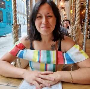 A woman with shoulder-length dark hair, wearing a colourful striped top, sits in a cafe, smiling at the camera.