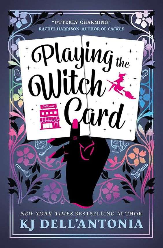 The front cover for Playing the Witch Card by K.J Dell'Antonio. The front cover is shades of purple. There is a black hand with pink fingernails rising out of the bottom of the page. The hand is holding a pair of white cards with the book's title running across them. There is a small image of a pink witch on a broomstick on the middle right of the title and a pink building to the bottom left of the title. Around the hand decorating the rest of the cover is a floral motif of flowers, stalks and leaves in shades of purples and pinks.