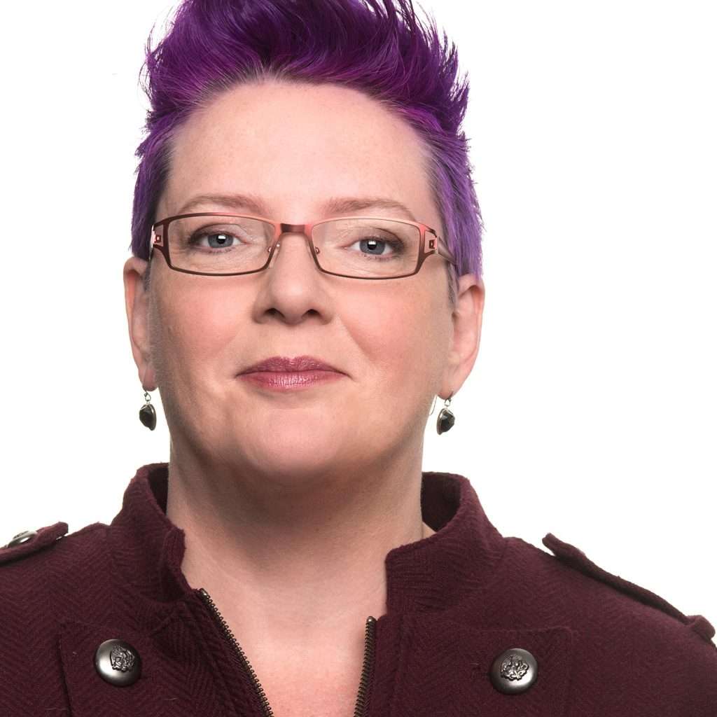 A person with short purple hair and glasses, wearing a maroon coat, looking towards the camera, against a white background. 