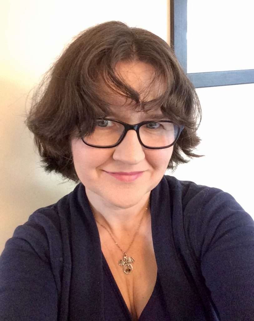 A woman with short, brown hair and black glasses, wearing a dark blue top, sitting in front of a window.