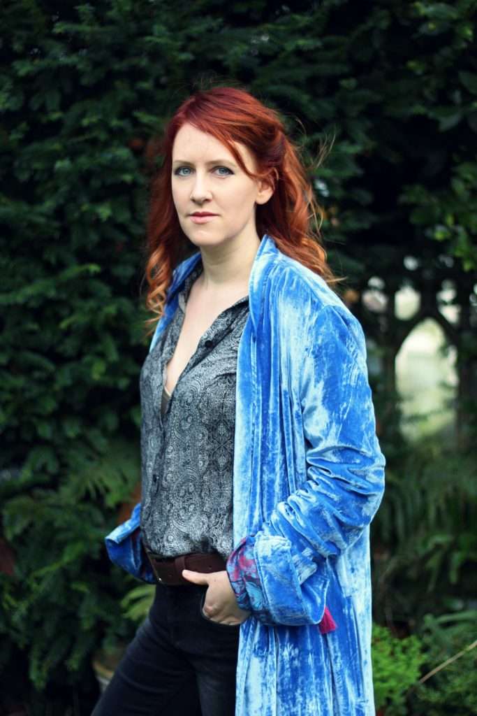 A woman with long red hair, wearing jeans, a blouse, and a long, blue velvet coat, standing in front of trees and bushes.