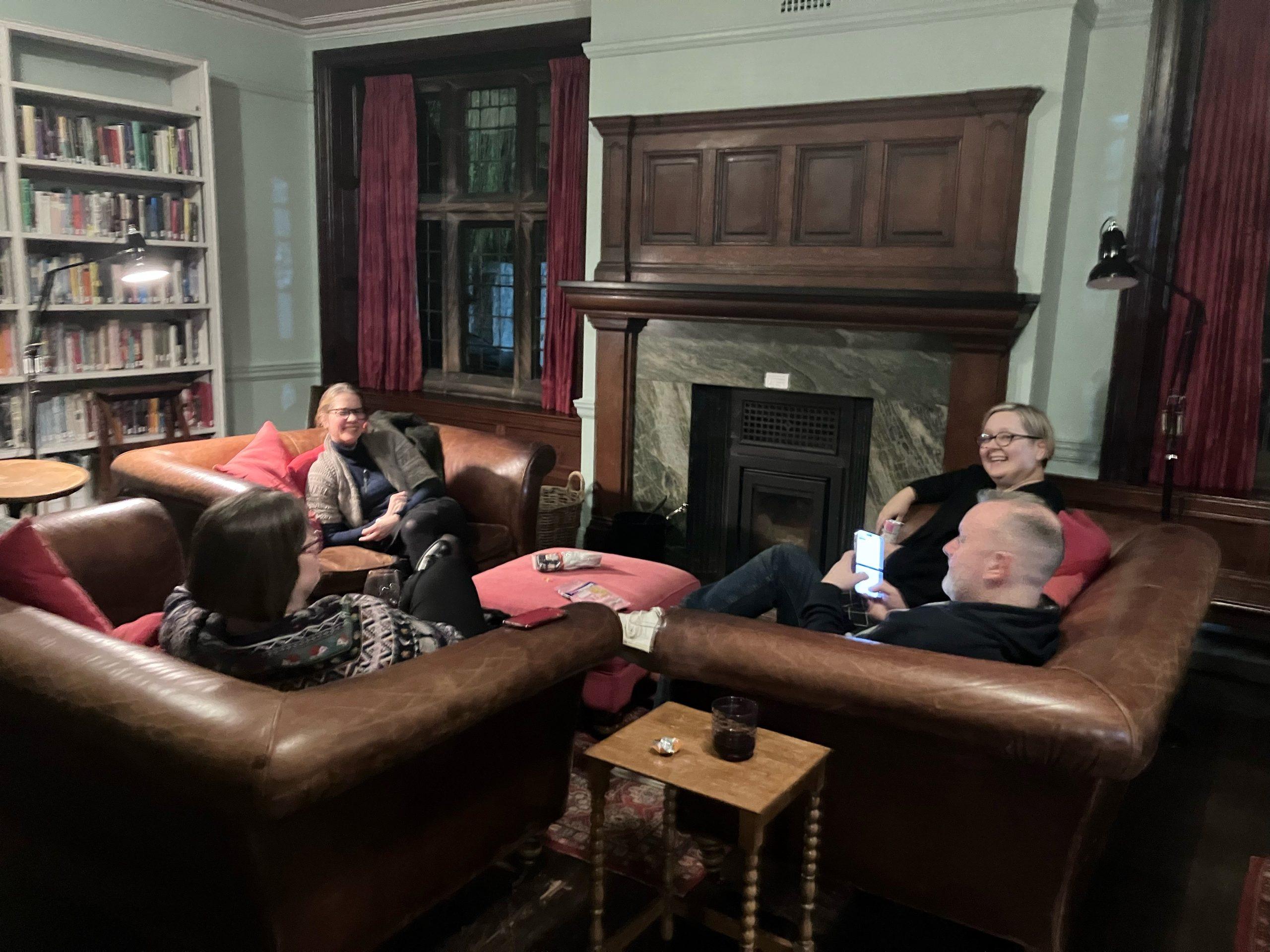 A group of people sit on leather sofas around a traditional fireplace. They are talking and laughing. Arched windows are on either side of the fireplace and bookcases line one wall. 