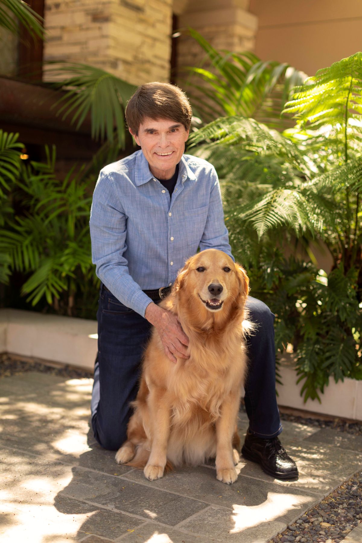Dean Koontz is wearing a light blue shirt and dark blue trousers, standing behind his golden retriever, Elsa.