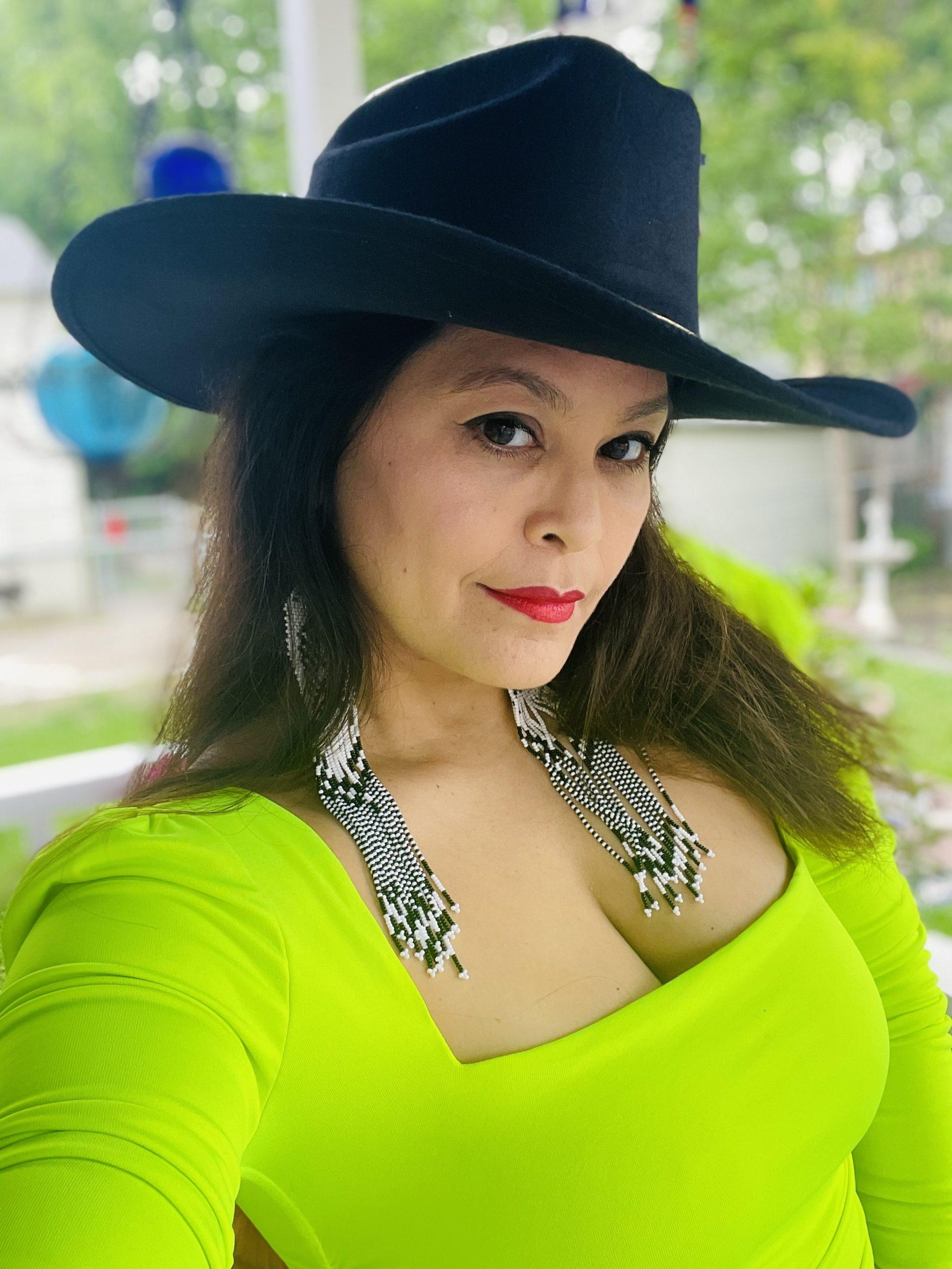 An image of a Mexican American woman smiling towards the camera. She has long, dark hair and is wearing a black stetson hat and lime yellow/green top