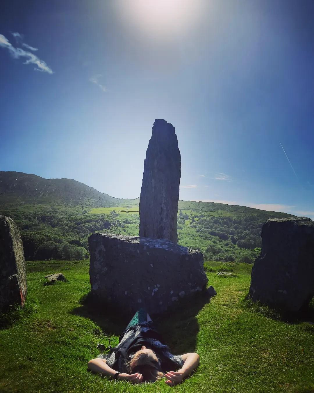 Tracy Fahey lies on the ground in front of a monolith in the midst of green fields. She's at the Uragh Stone Circle while doing fieldwork in Ireland.