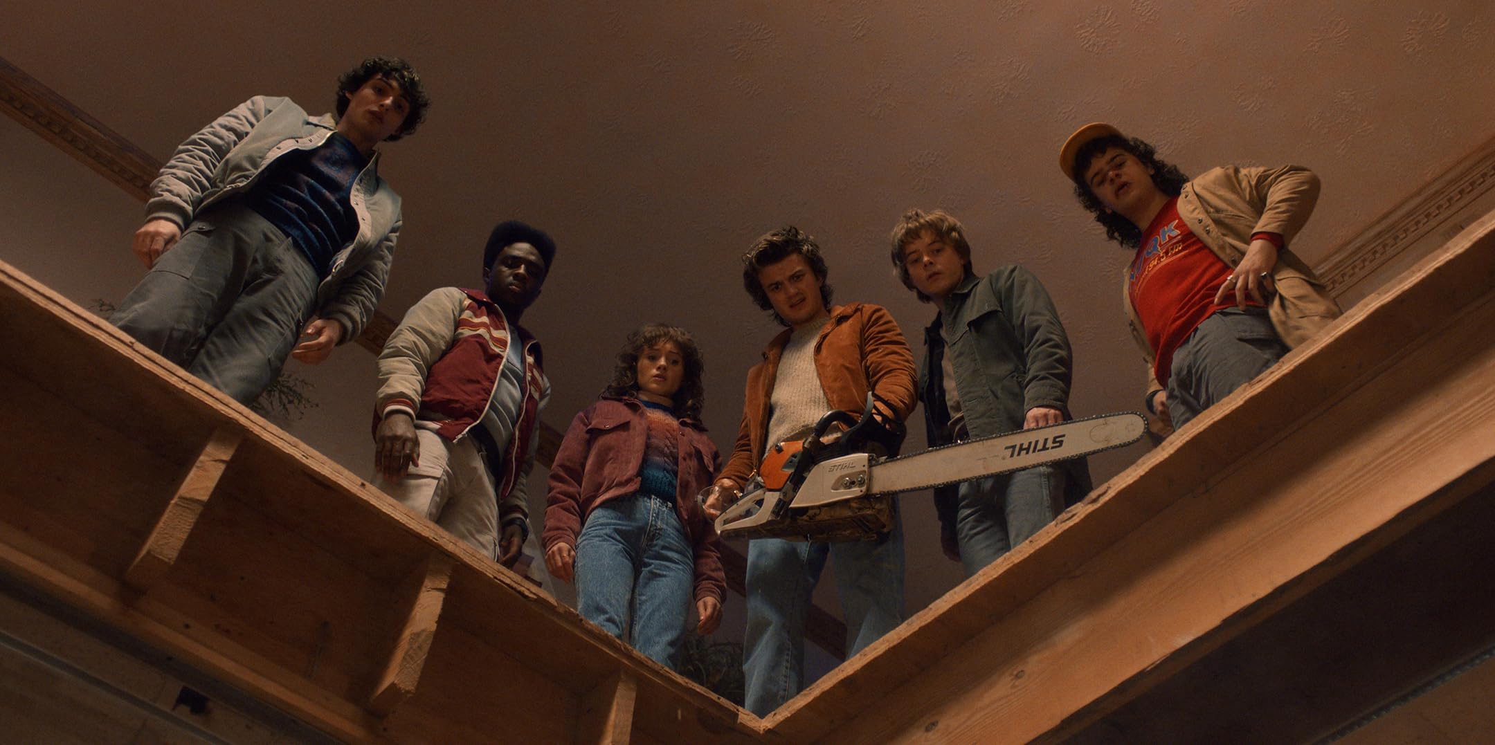 The cast of Stranger Things with a chainsaw look down at the camera from a walkway