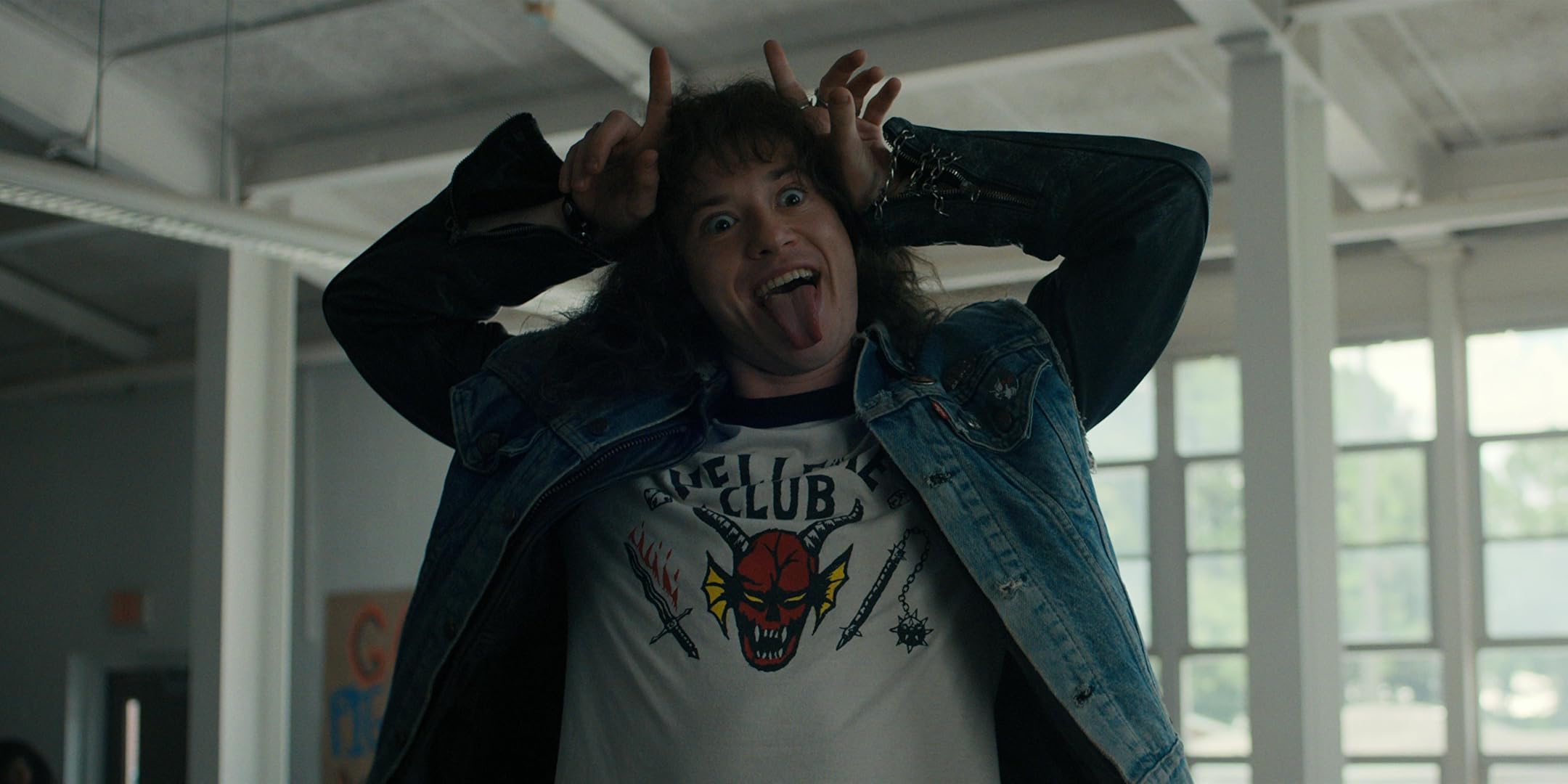 A man with an 80s metal fan haircut pulls a face and holds his hands to his head as if they were horns