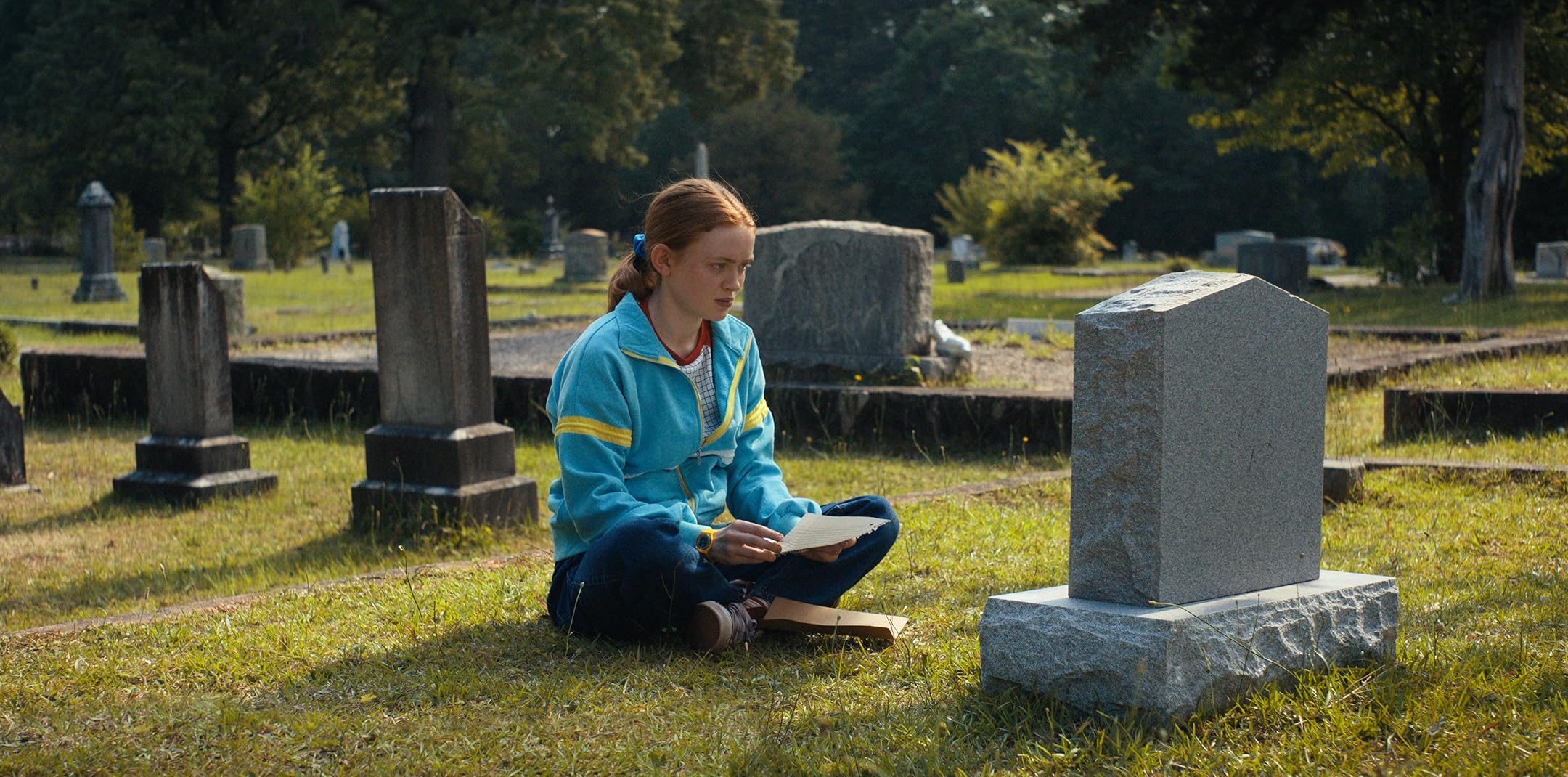A girl sits cross legged in a graveyard, facing the grave
