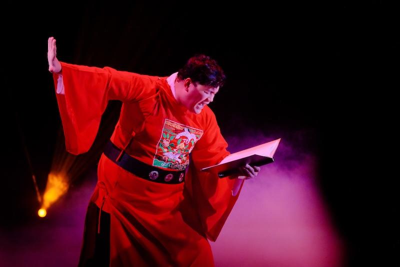 A scene from Asian Pirate Musical shows a performer in traditional Chinese dress singing while reading a book