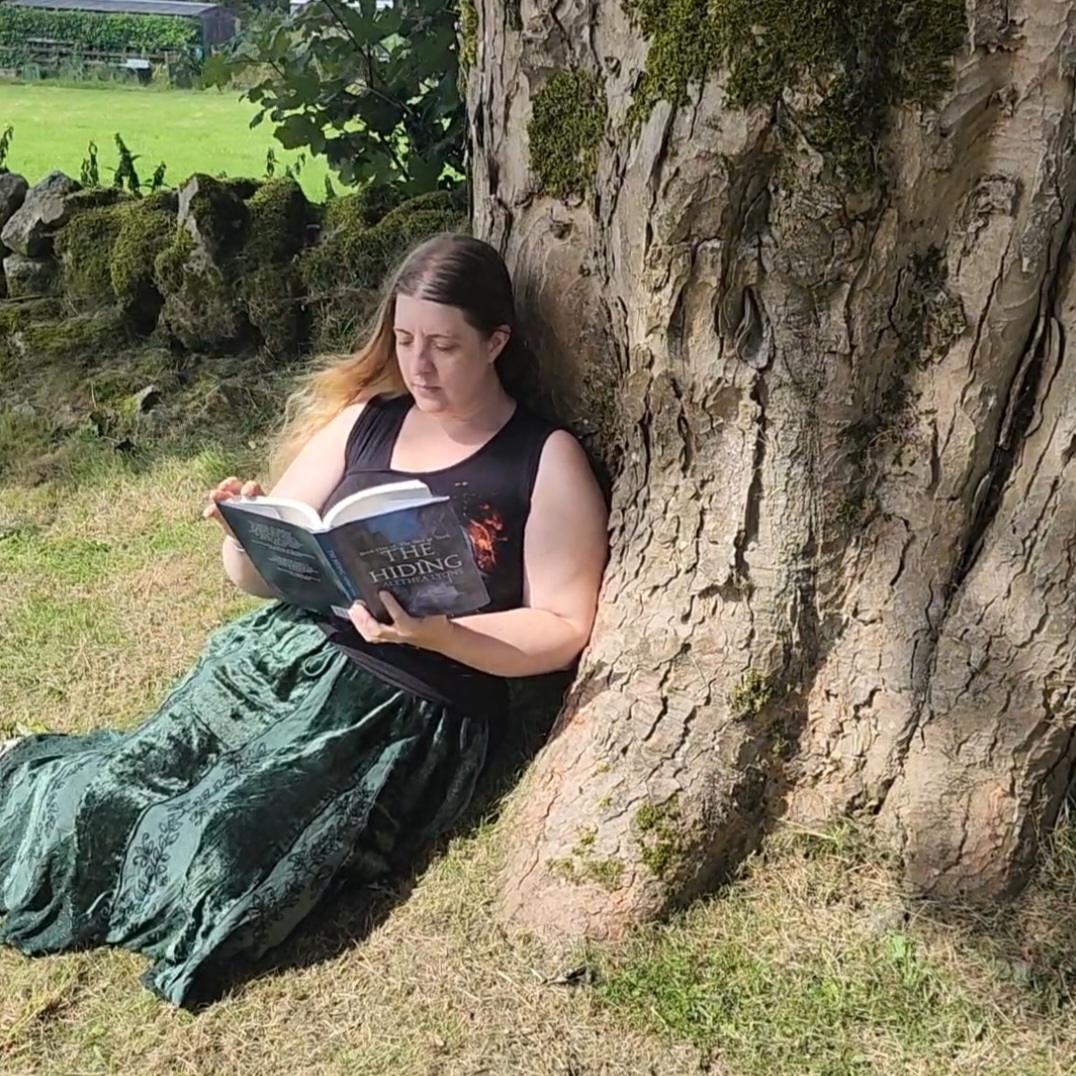 Author Alethea Lyons sits on the ground, leaning against a tree, reading a book