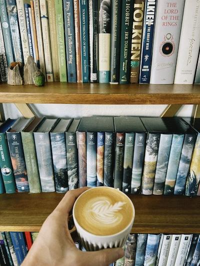 A hand holds a coffee up in front of bookshelves full of classic fantasy novels