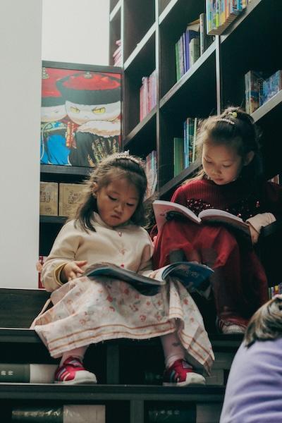 Two young East Asian girls sit on stairs in a library, reading books