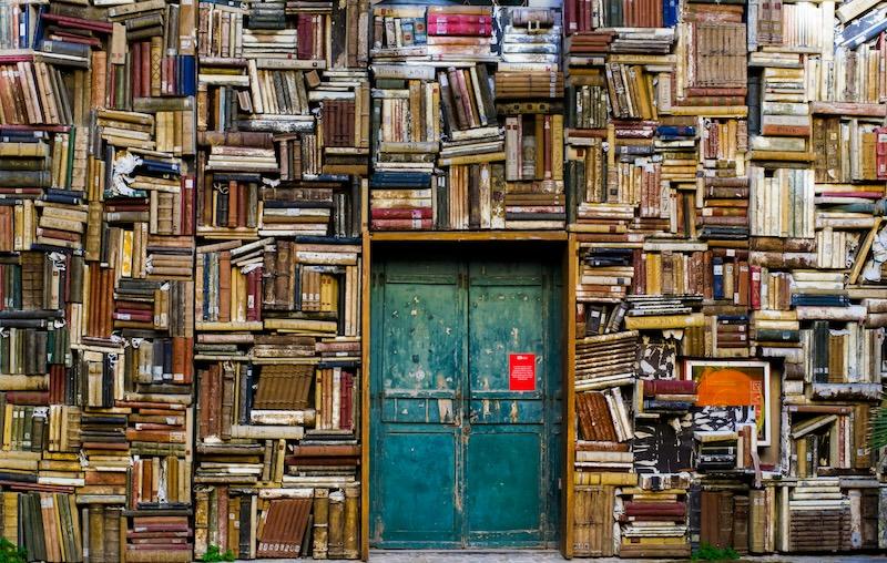 A wall full of bookshelves overflowing with old books. There is a greenish-blue worn door in the middle. 