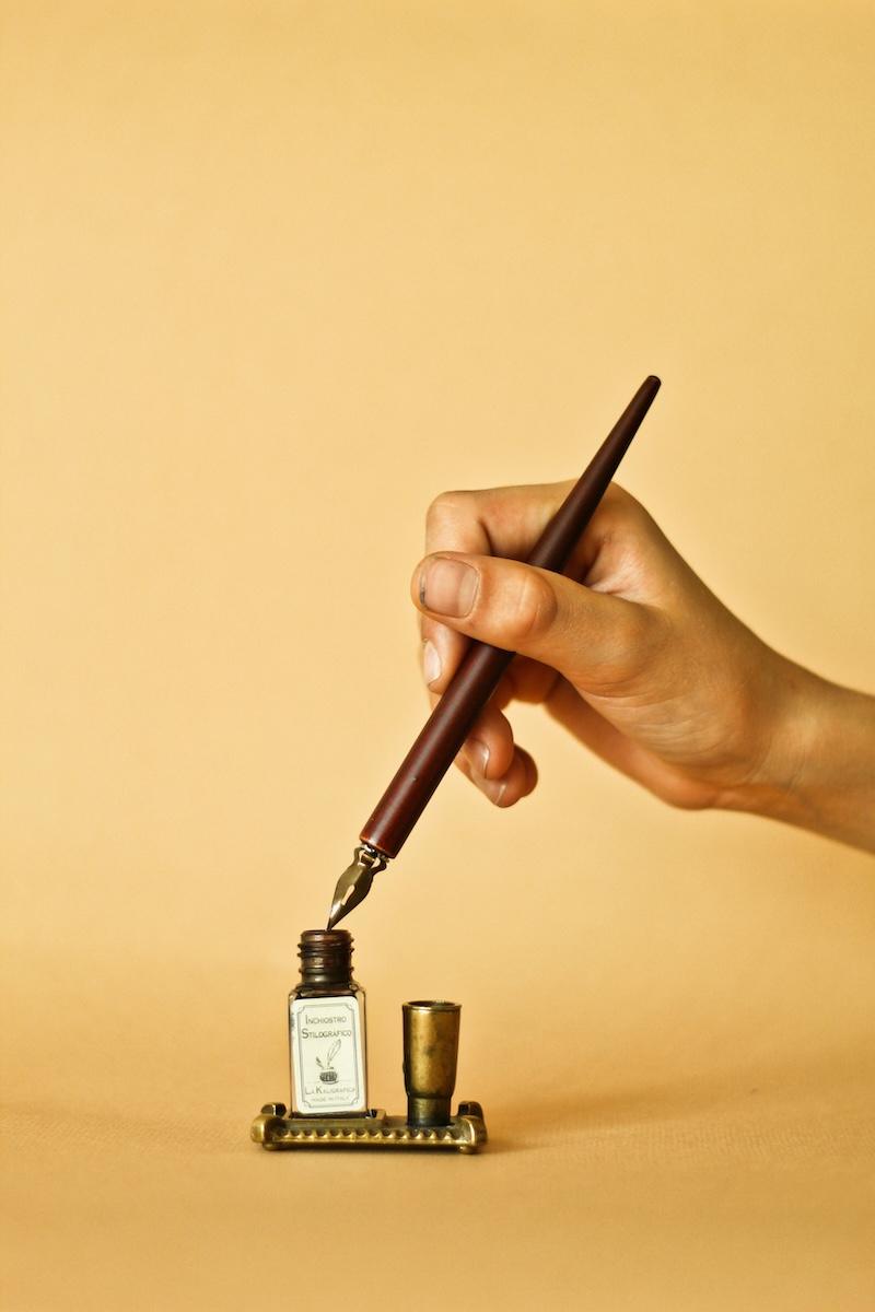 A hand touches a pen nib to a bottle of ink