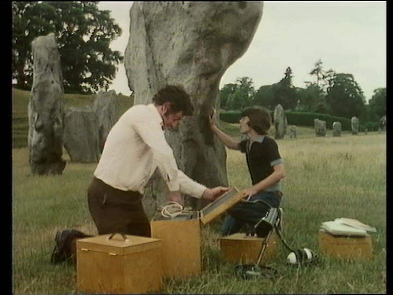 A man and a boy unpacking technical equipment in a British stone circle. The boy is touching one of the megaliths.