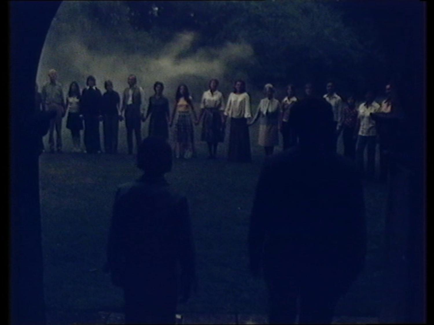 A row of people standing holding hands. We are looking from behind two people. The image is vaguely sinister, it is at night.