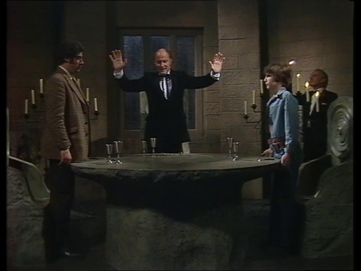 People stand around an old table. A man has his hands raised. There are goblets on the table, and candleabra behind, as if a ritual is about to be performed.