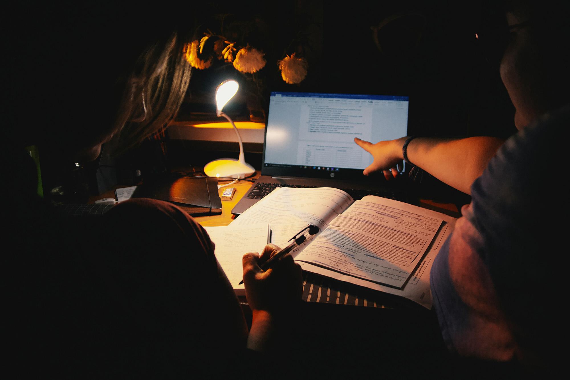 A darkened room with a small light, two people are working in front of a laptop, one is pointing at the screen, the other is making notes.
