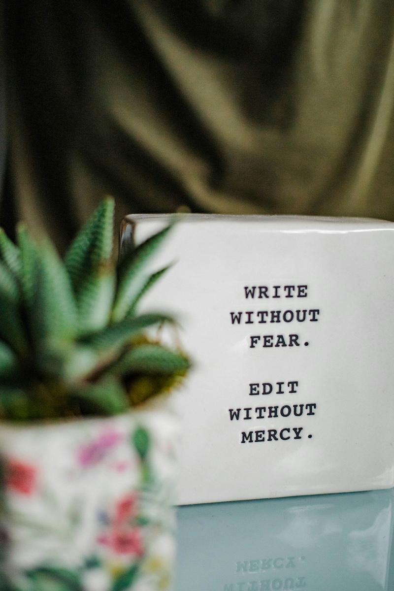A white box has the text "write without fear; edit without mercy". It sits on a table next to a plant.