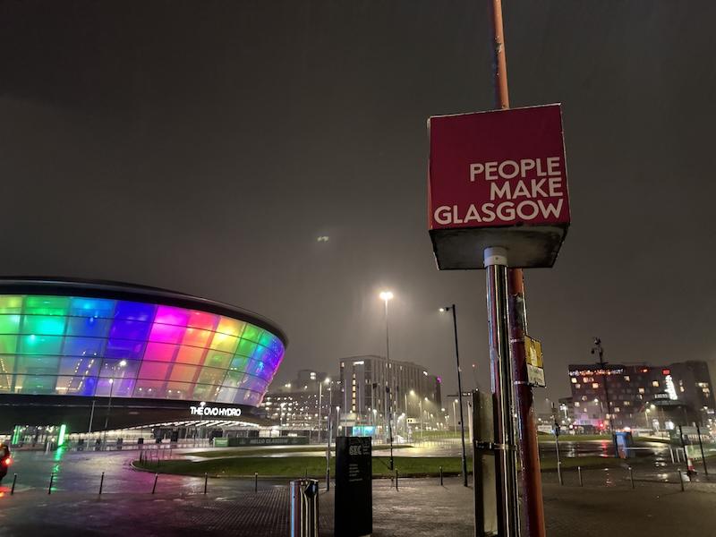 The Glasgow Ovo Hydro stadium lit in rainbow colours, with a sign saying "People Make Glasgow" in the foreground